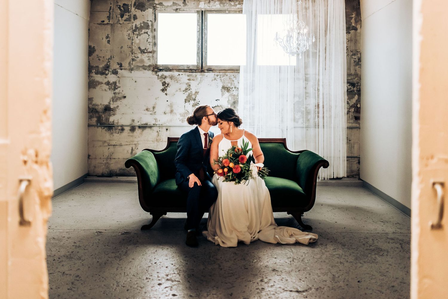 Intimate wedding portrait on an emerald velvet sofa in an industrial-style room with distressed walls.