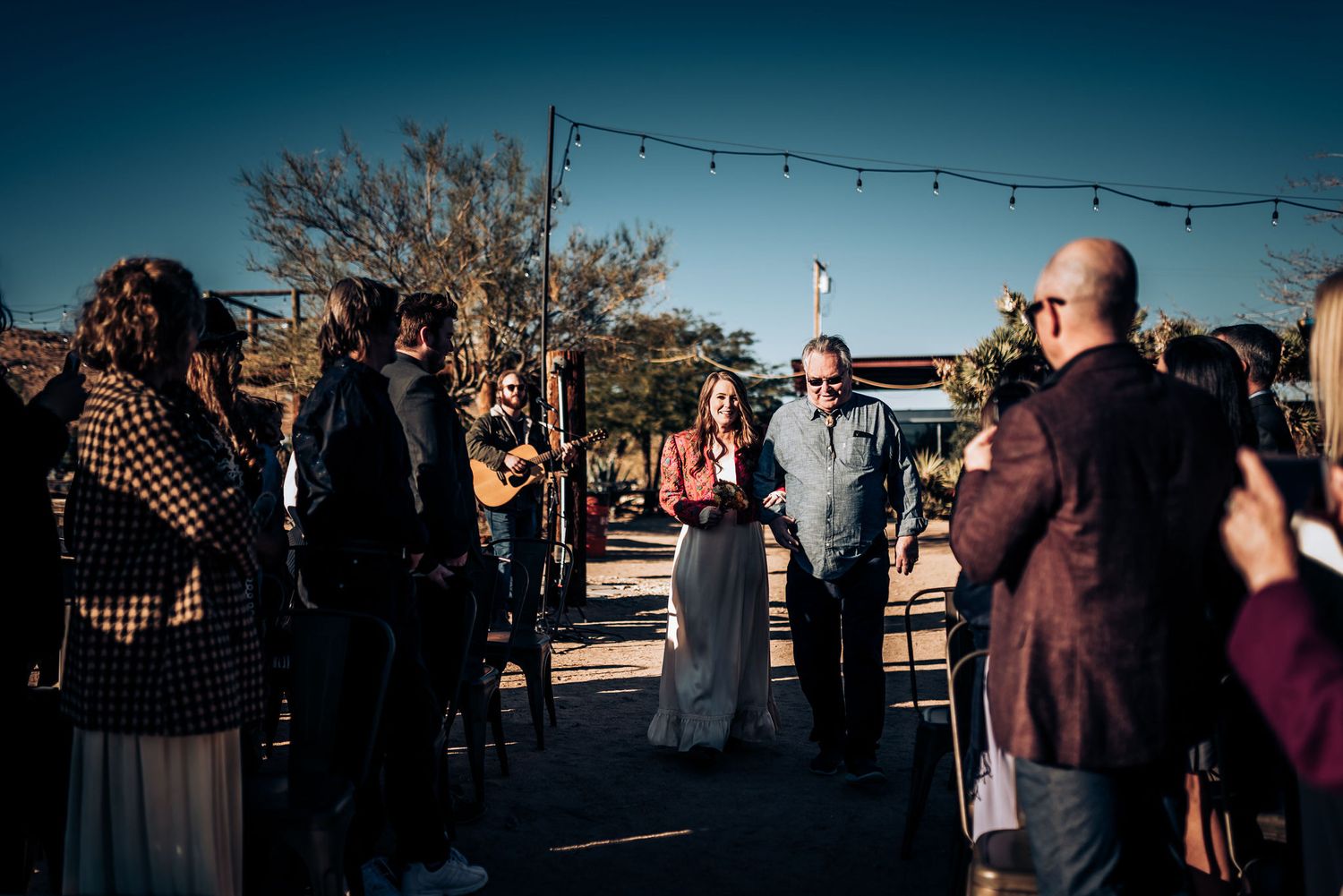 An outdoor gathering with string lights overhead as guests stand together at a desert venue during sunset.