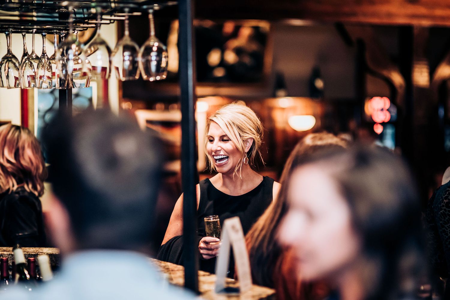 People gather at a warmly lit bar with wine glasses hanging overhead during a celebration.