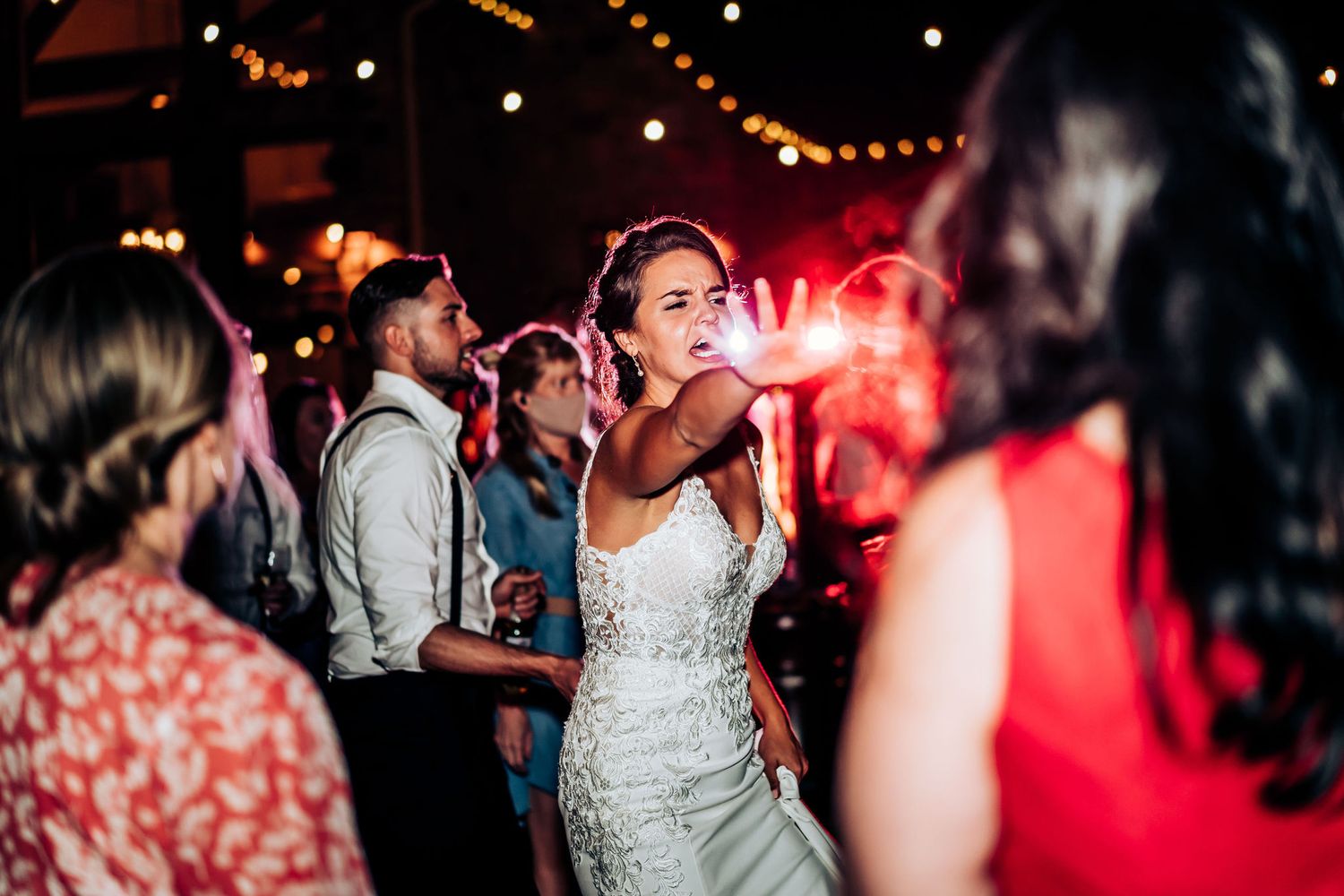 A wedding reception party in full swing with dancing under string lights and red ambient lighting.