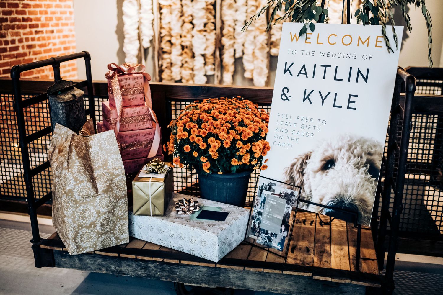 Wedding welcome sign display with orange flowers and decorative vase on brick backdrop.