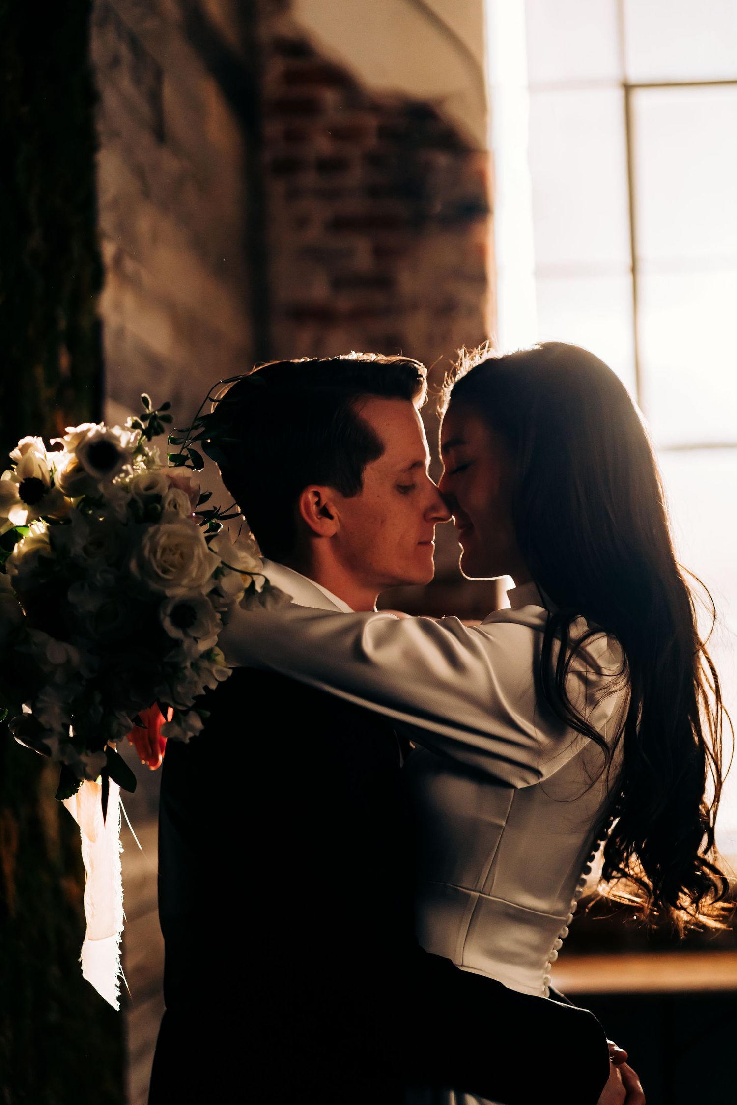 Silhouetted couple sharing an intimate moment in front of sunlit windows.