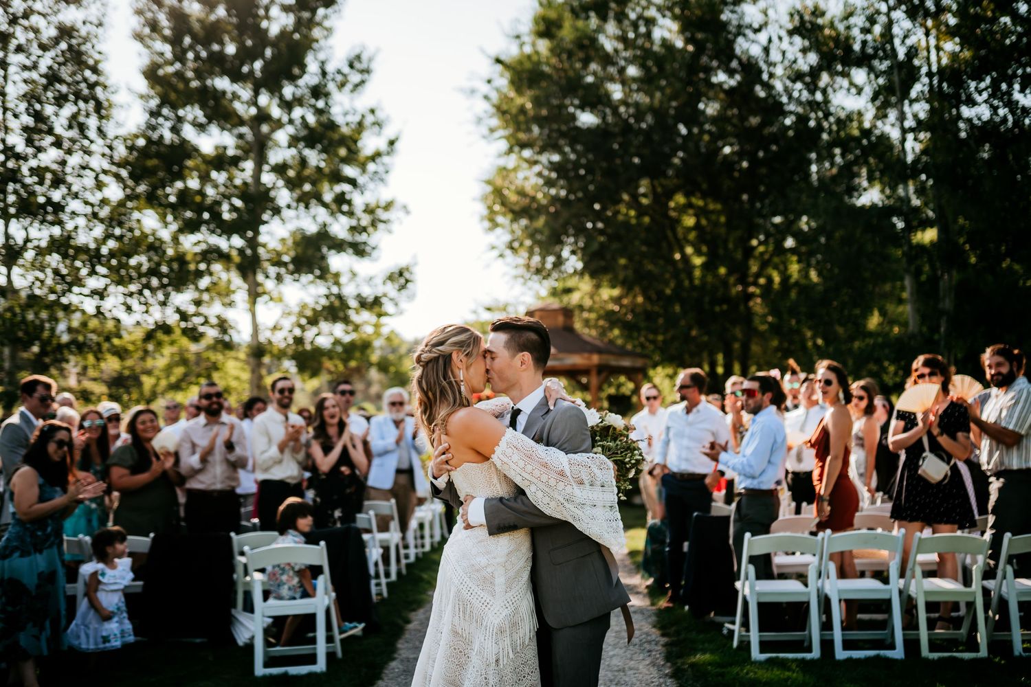 Outdoor wedding ceremony with guests seated on white chairs at sunset.