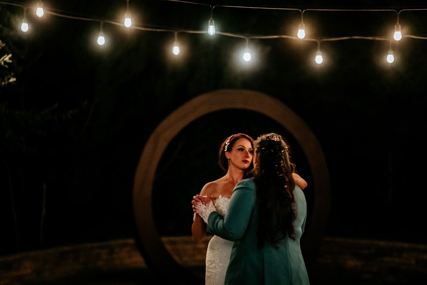 Two brides share a romantic dance outdoors at night beneath glowing string lights.