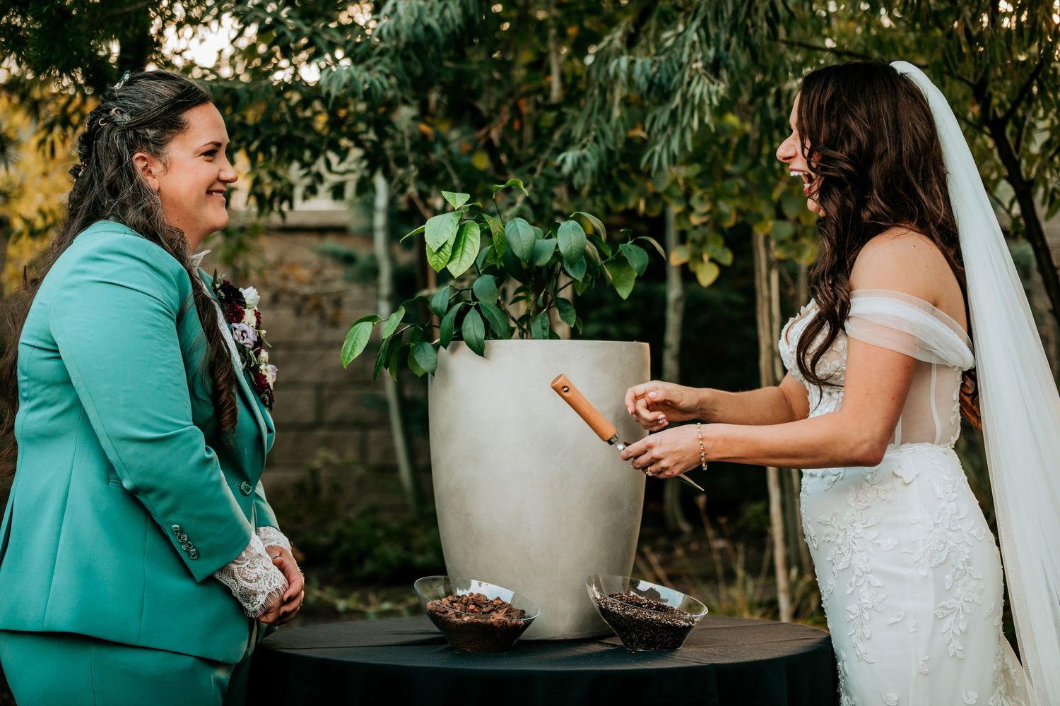 Two brides share a joyful moment during a unity plant ceremony outdoors, one in teal suit and one in lace gown.