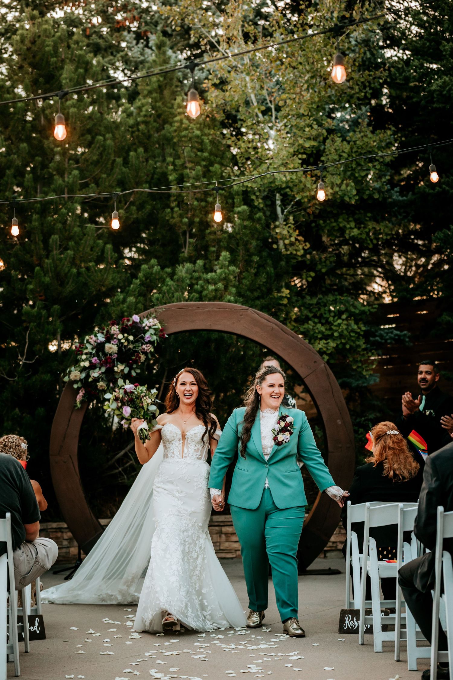 Two brides walk down a petal-lined aisle, one in a white gown and one in a teal suit, at their wedding.