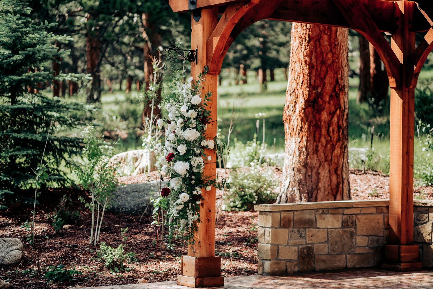 Stone and timber wedding pergola with floral decorations nestled among pine trees.