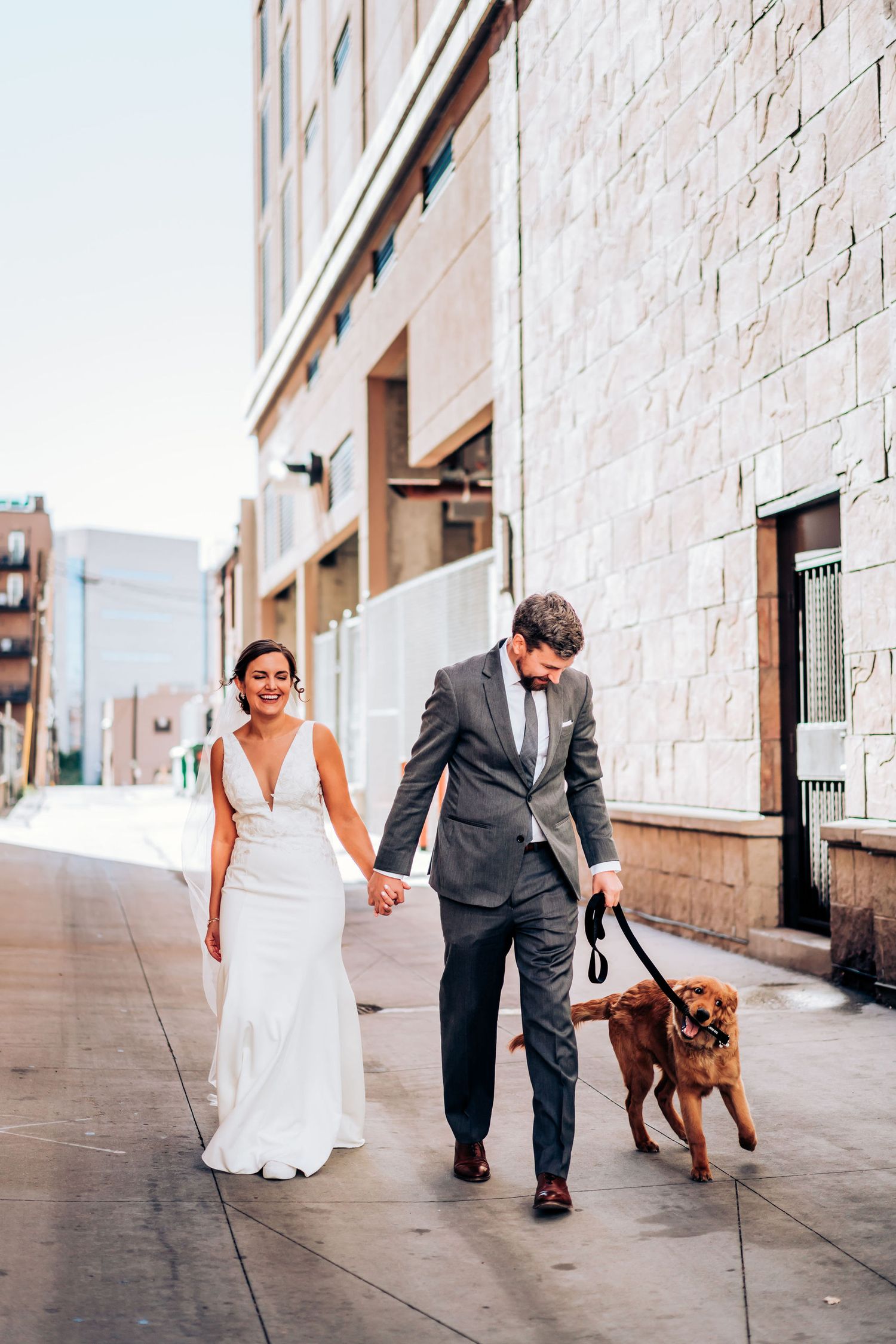 Wedding couple strolls down a city sidewalk with their brown dog on a leash.
