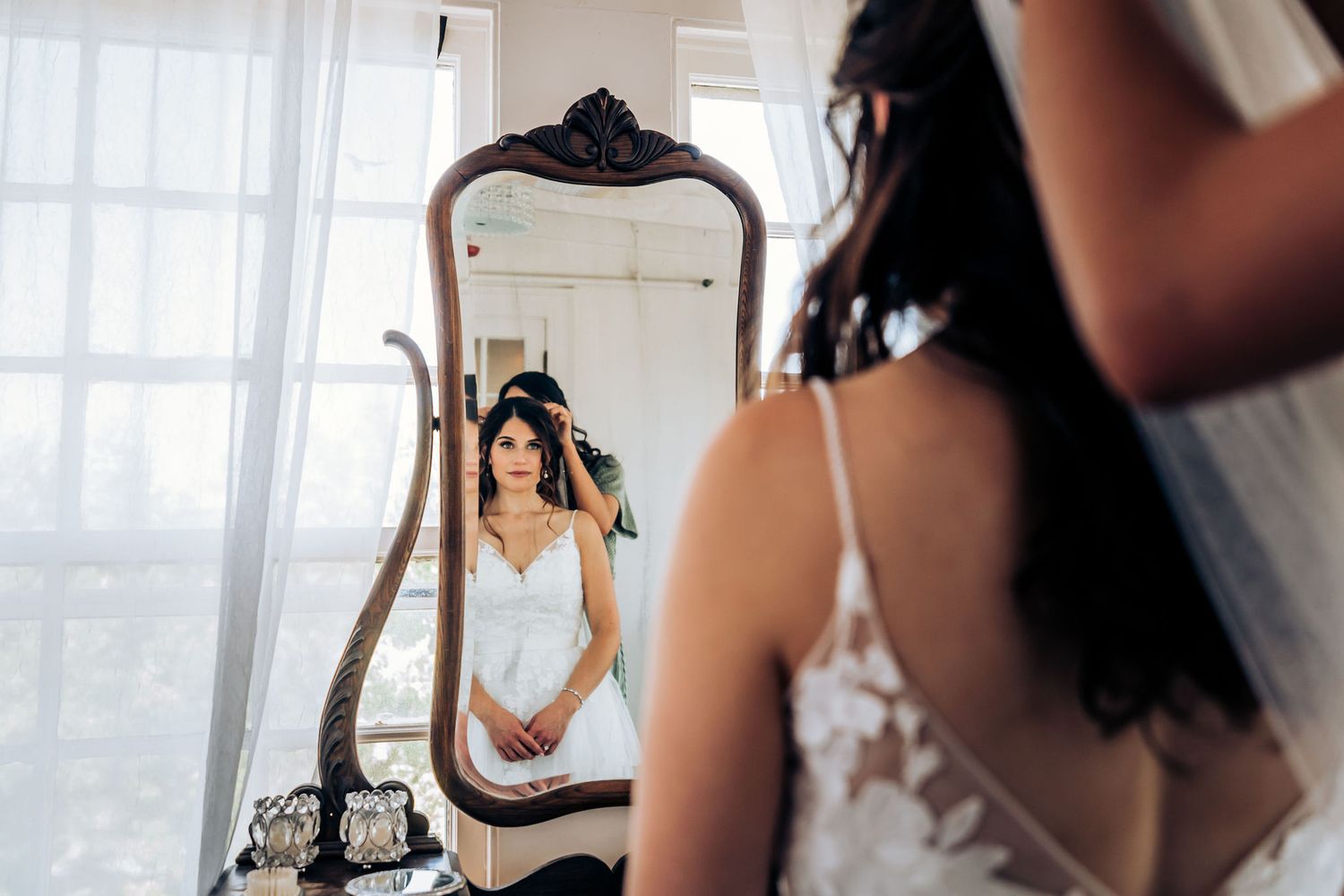 Elegant bride in white dress looking at her reflection in an ornate vintage mirror during wedding preparations.