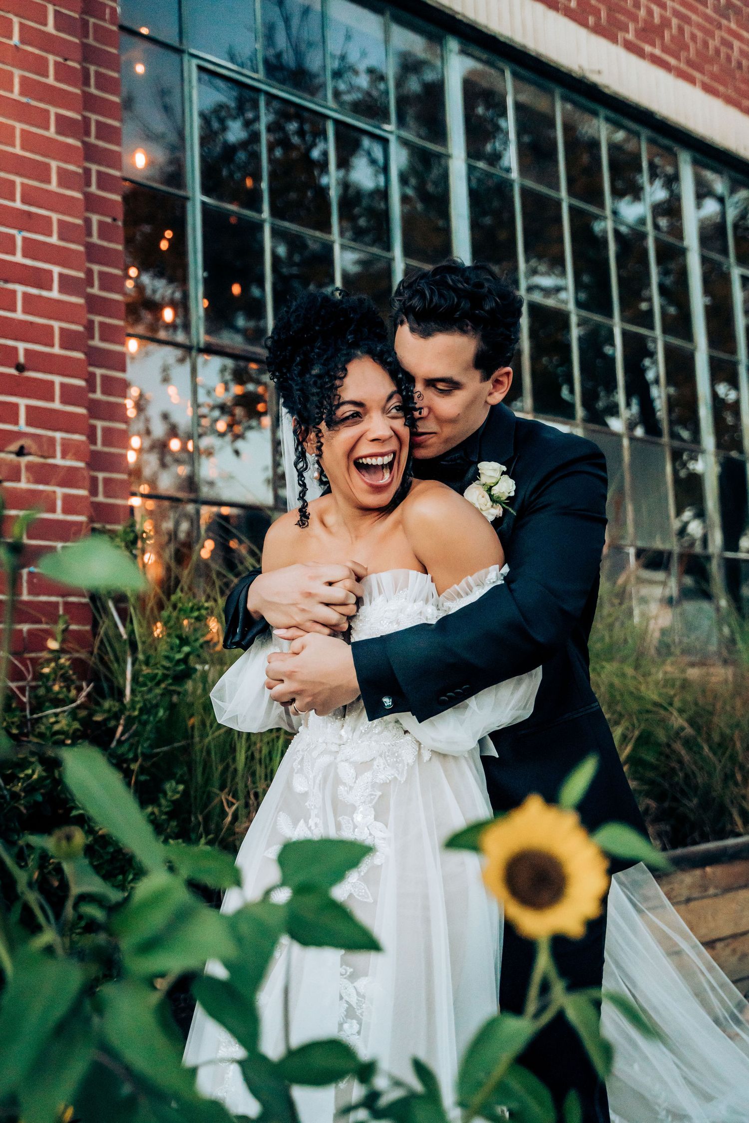 Joyful wedding moment captured in front of a brick building with sunflowers and string lights.
