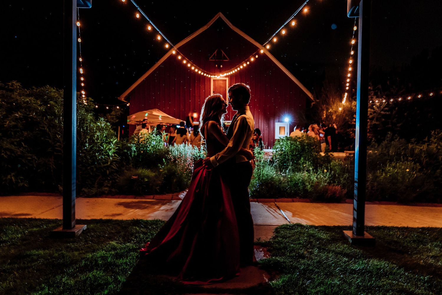 Romantic nighttime dance under twinkling lights and red neon cross installation in garden setting.