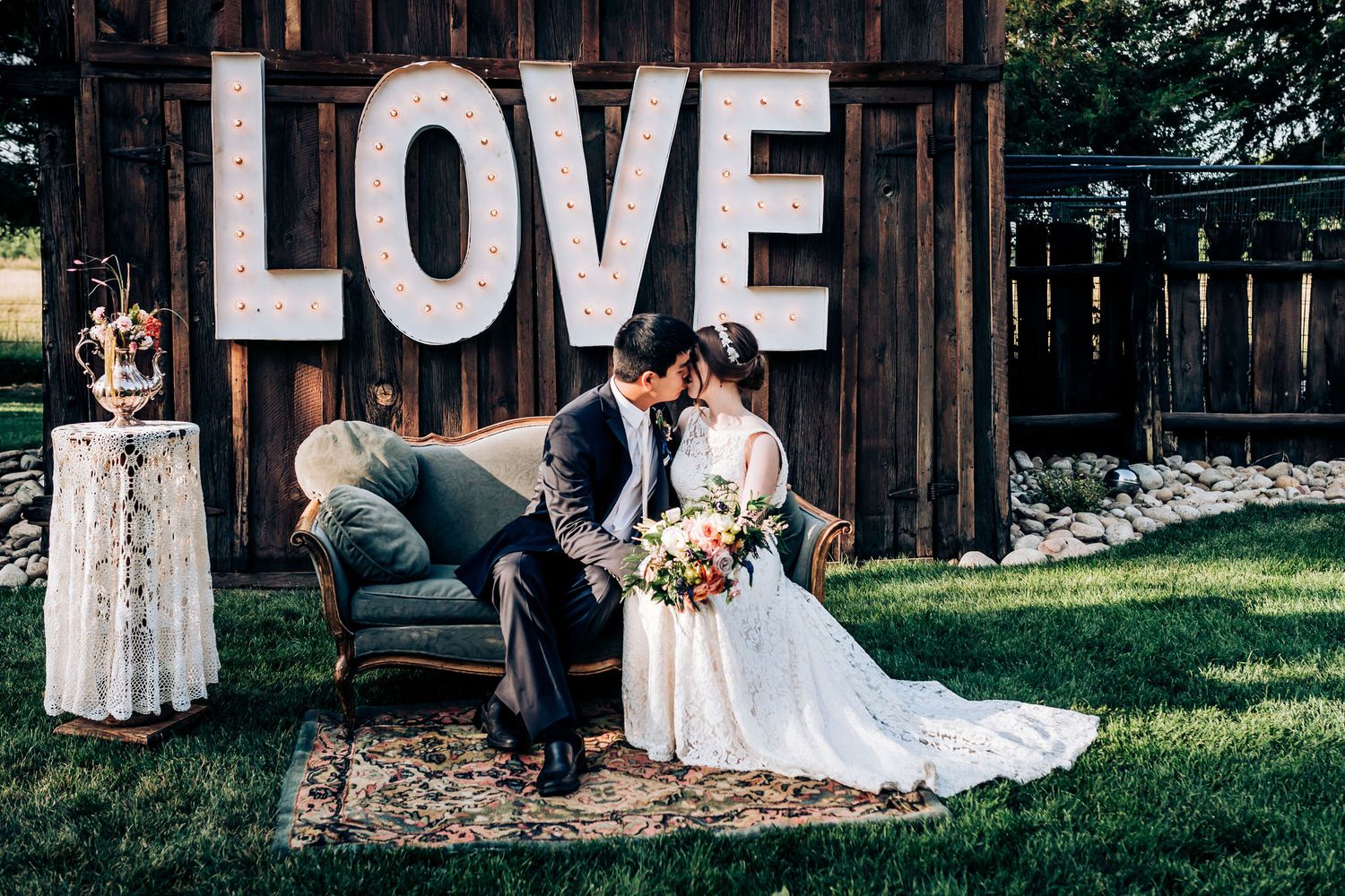 Wedding couple seated on vintage couch against rustic wooden wall with illuminated LOVE sign.