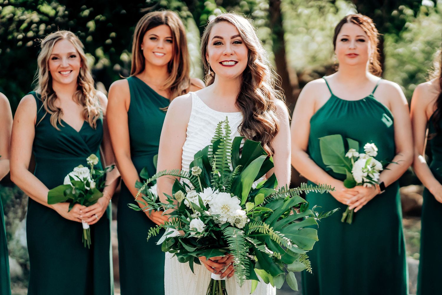 A bridal party poses outdoors wearing emerald green dresses and holding white floral bouquets.