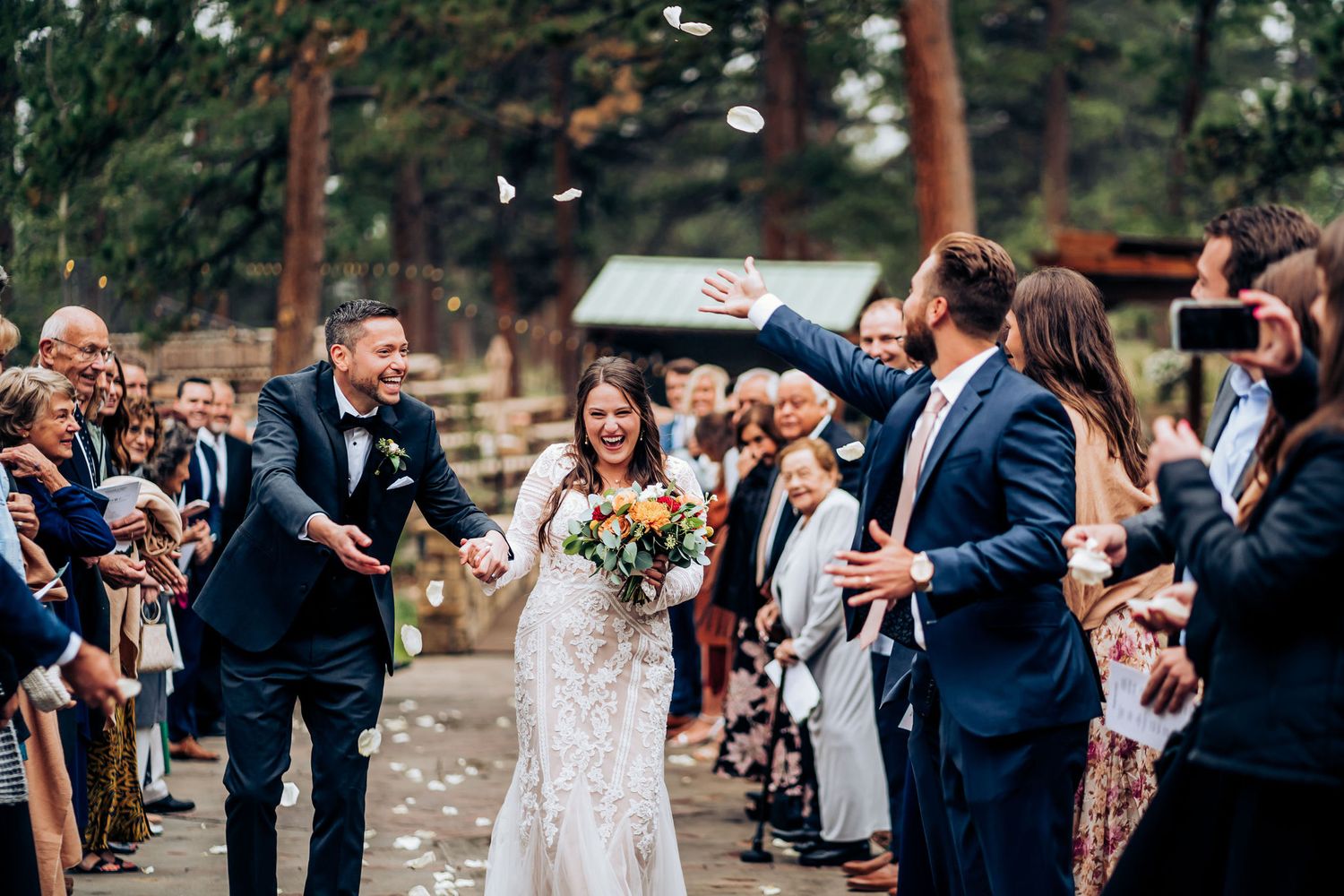 Wedding guests throw confetti as newlyweds walk down an outdoor ceremony aisle.