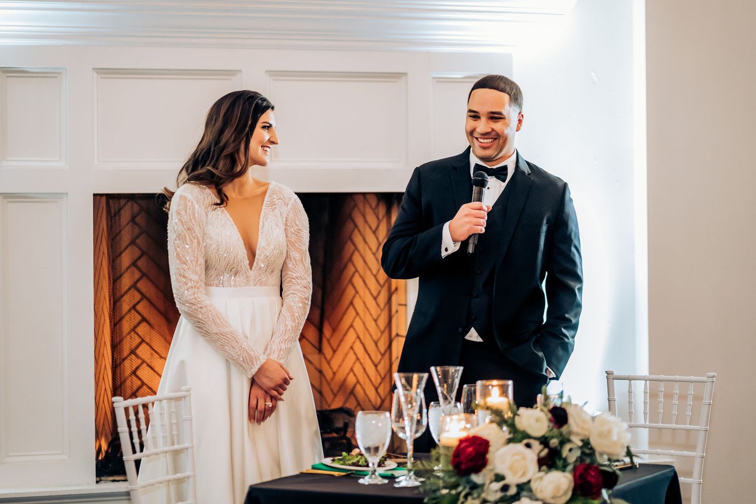 Couple gives wedding reception speech at decorated head table with fireplace backdrop.