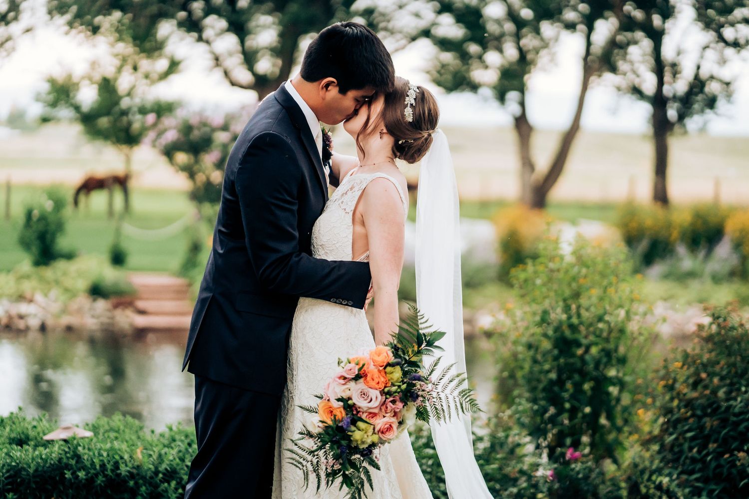 Romantic kiss between newlyweds by a pond surrounded by lush greenery.