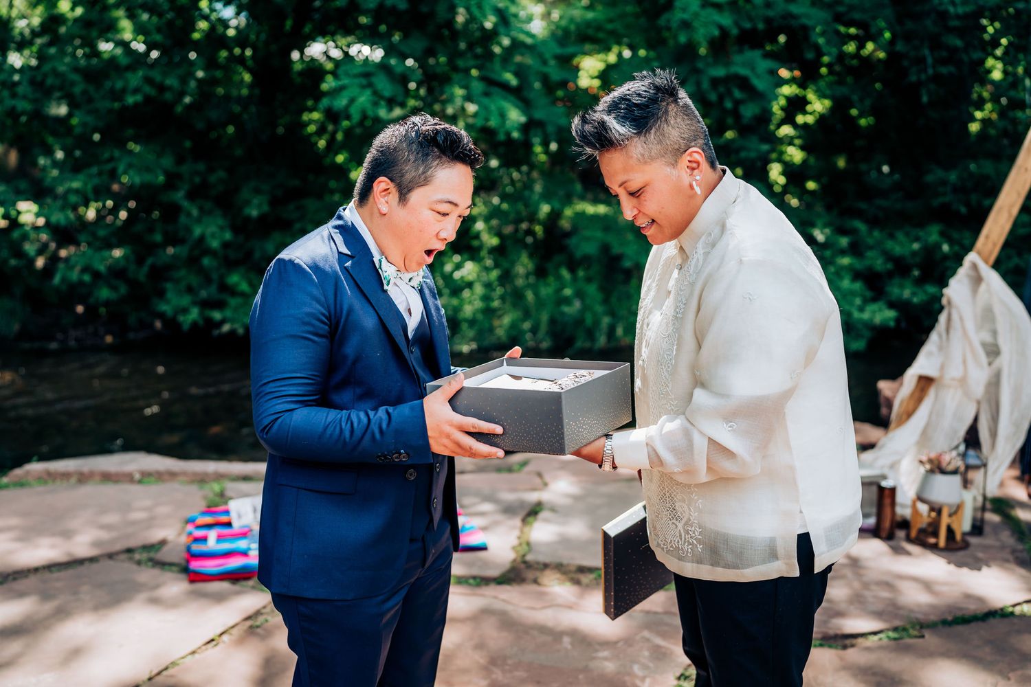 Two people exchange a gift box at an outdoor event, one wearing a navy suit and one in cream formal wear.