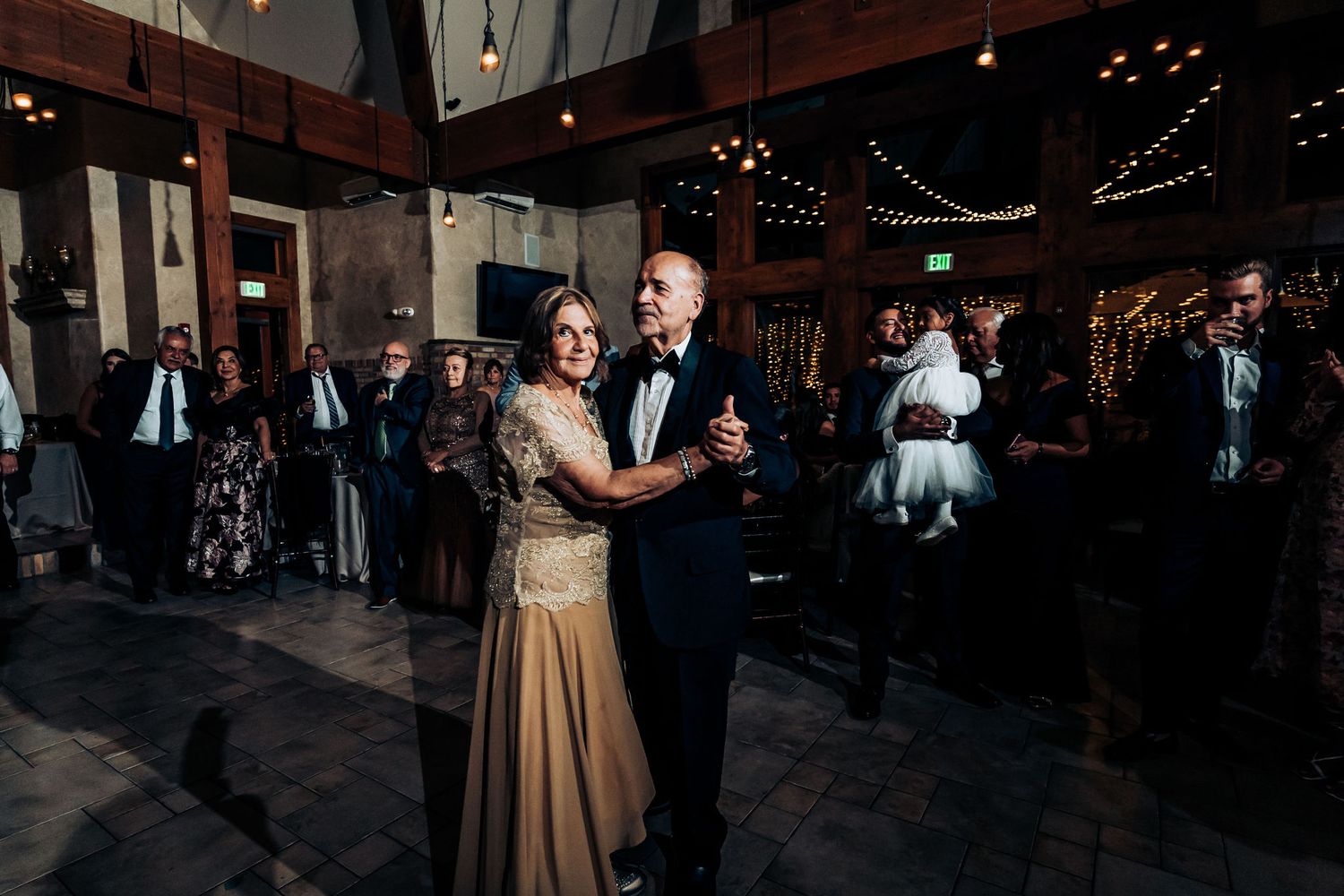 Elegant couples slow dancing in formal attire at wedding reception in rustic barn venue with wooden beams.