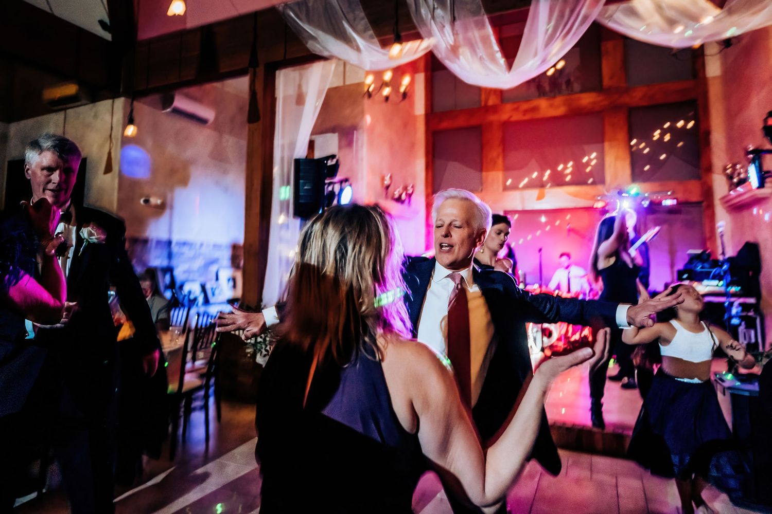 Wedding guests enjoying dancing and celebrating in a venue with pink ambient lighting.