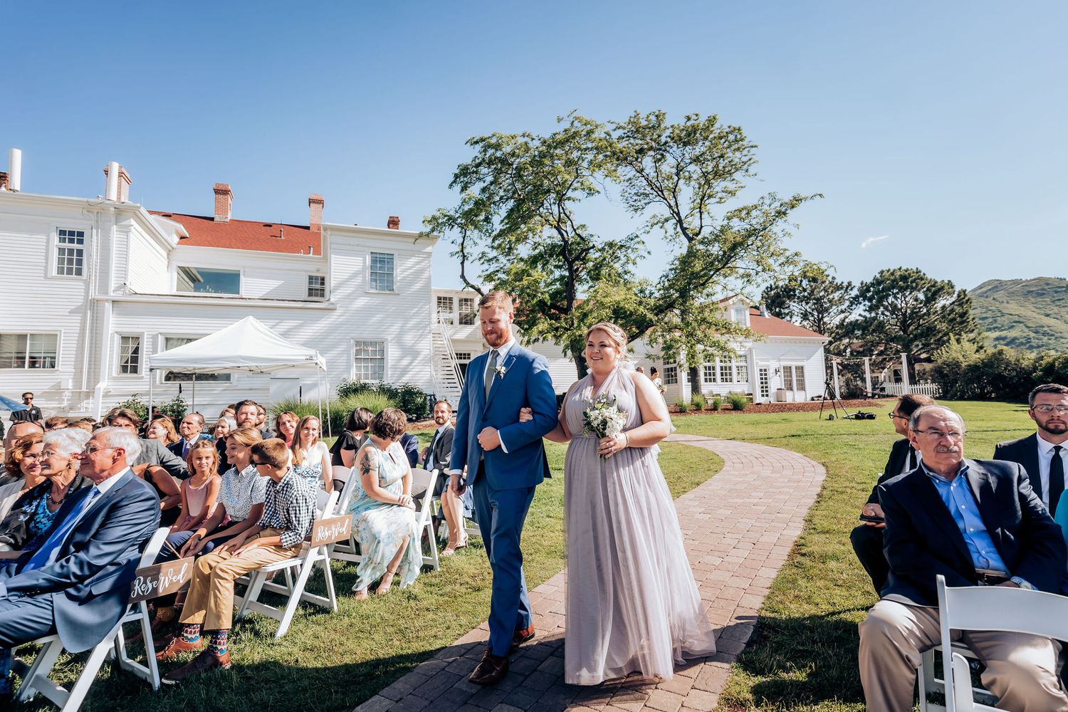 Wedding processional on brick path with guests seated in white chairs against mansion backdrop.