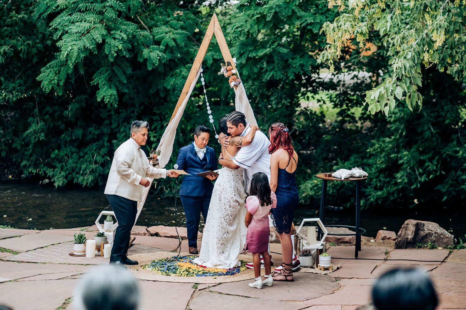 Wedding ceremony under a wooden triangle arch with white fabric draping in a natural outdoor setting.