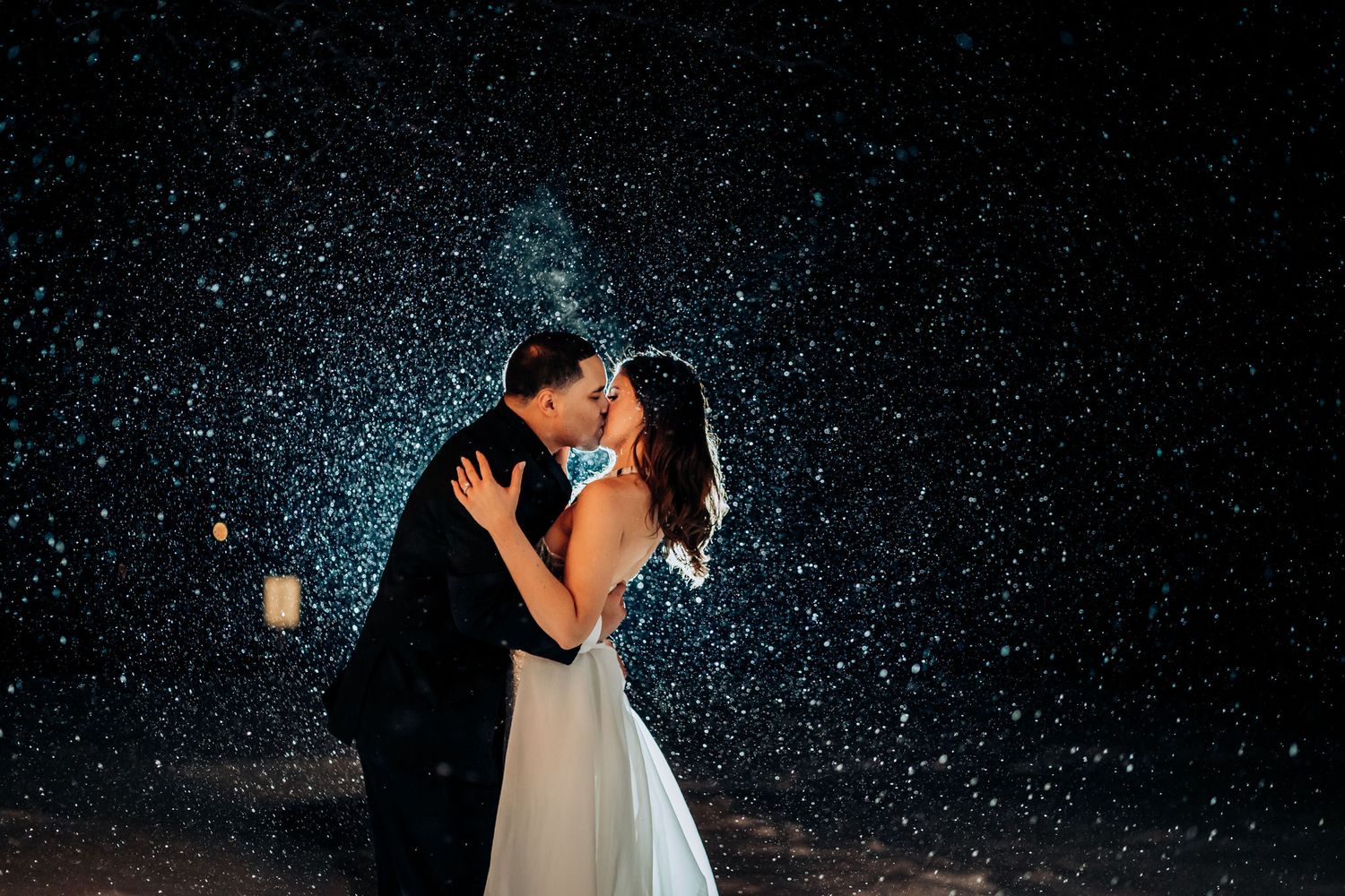 A romantic kiss under falling snow at night with twinkling lights in the background.