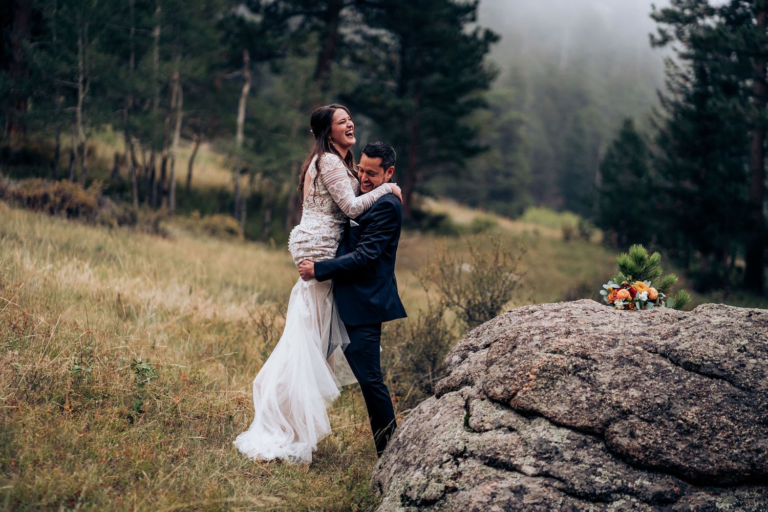 Wedding couple embraces on a large boulder in a misty mountain setting with pine trees.