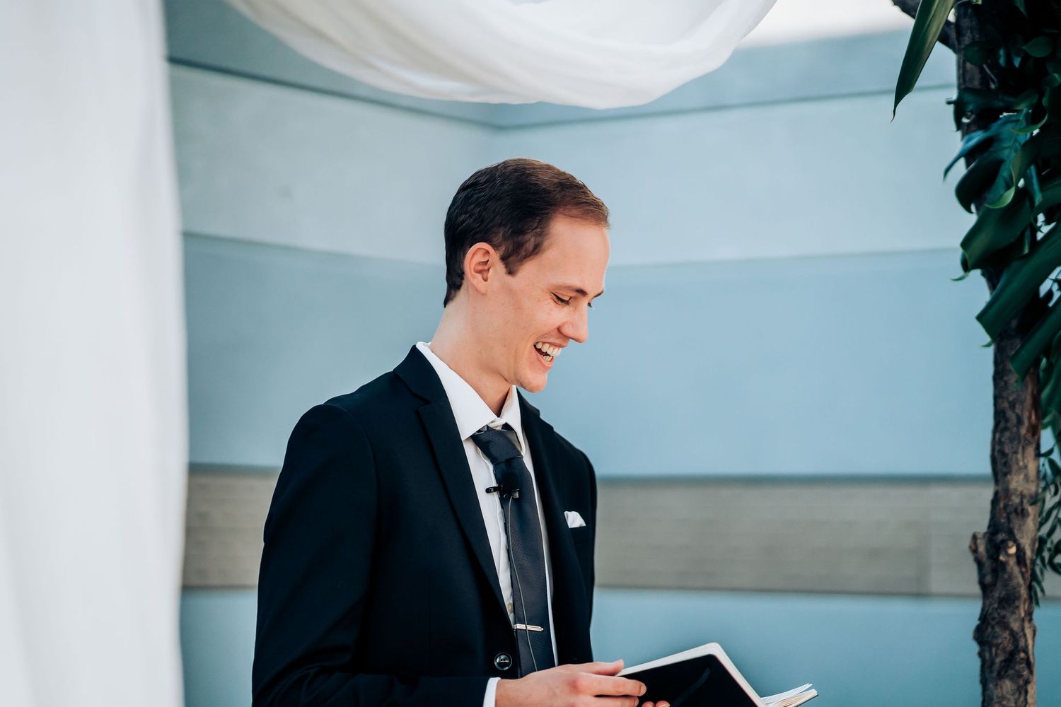 Wedding officiant in black suit reads from book during outdoor ceremony.