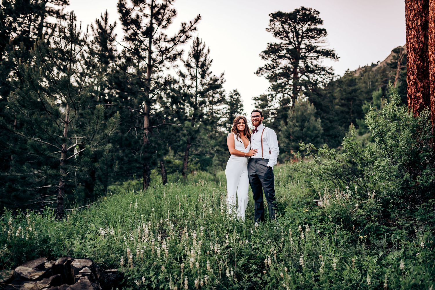 A couple embraces in formal attire against a backdrop of tall pine trees and mountain landscape.