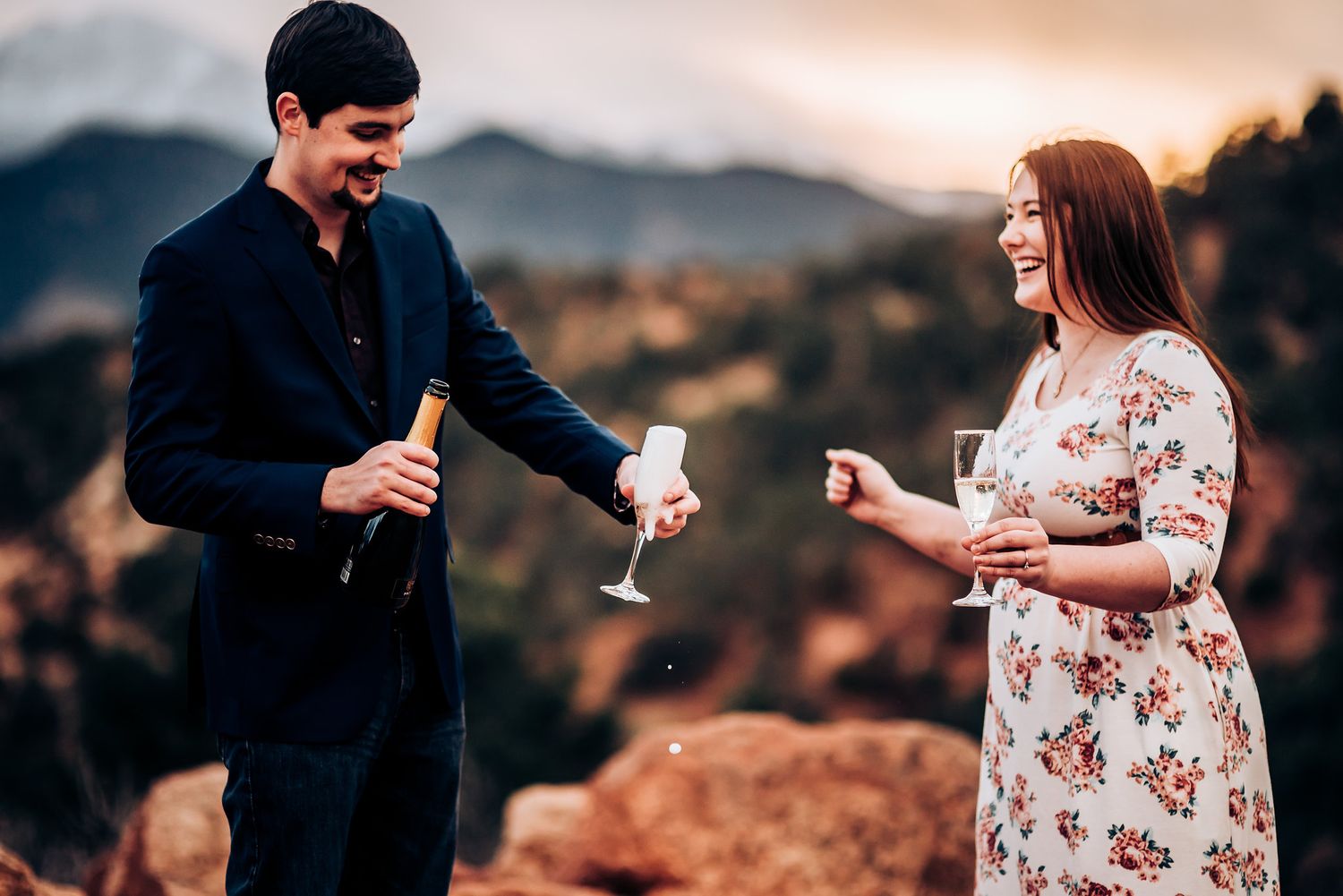 A couple laughs while pouring champagne on a scenic mountain overlook.