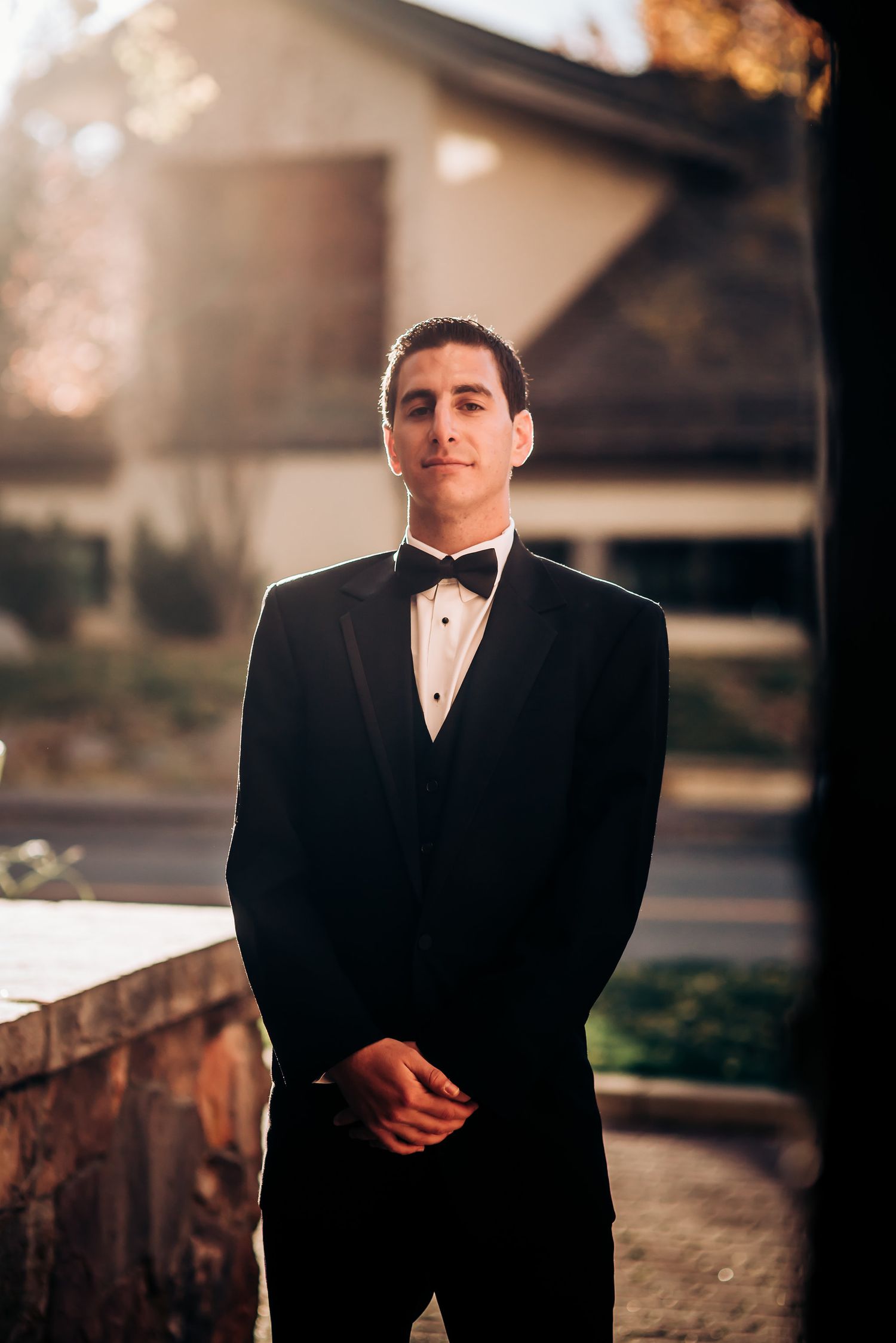 A formal portrait of a groom in classic black tuxedo standing on stone patio.