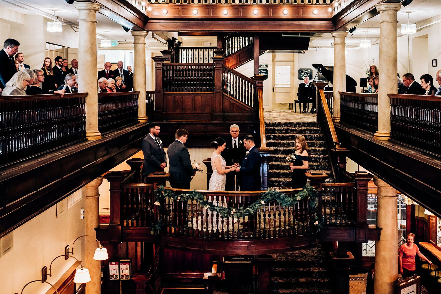 Wide view of wedding ceremony in historic venue with multiple levels and wooden architectural details.