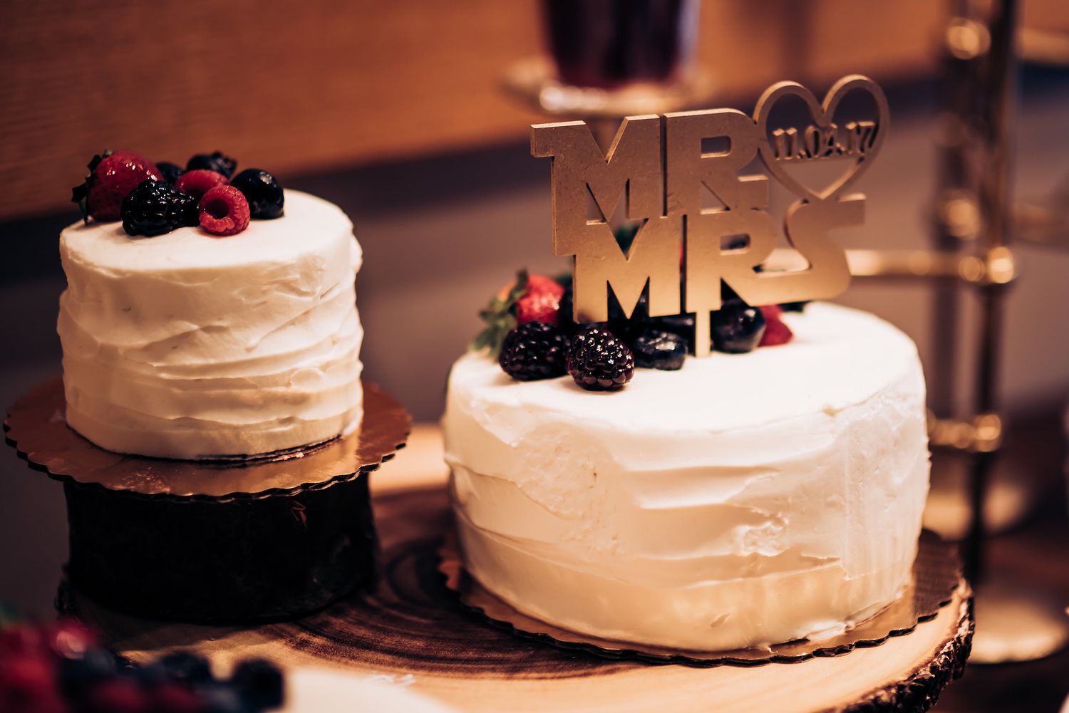 Two white wedding cakes decorated with fresh berries and a Mr & Mrs topper displayed on rustic wooden stands.