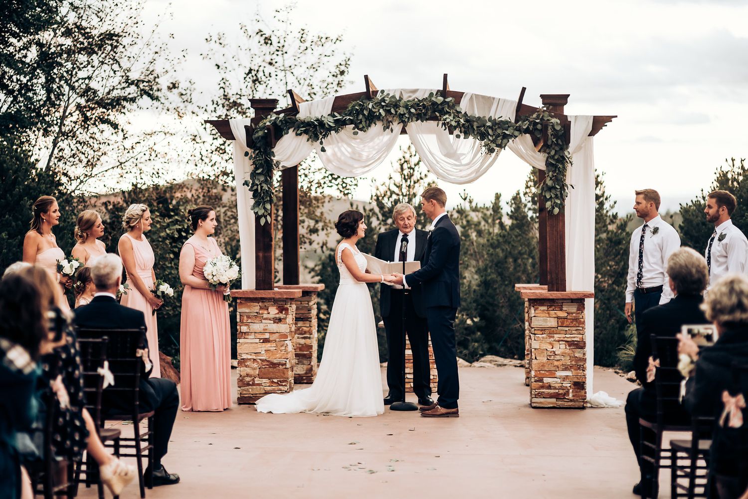 A wedding ceremony takes place under an elegant stone and fabric arch with guests seated outdoors.