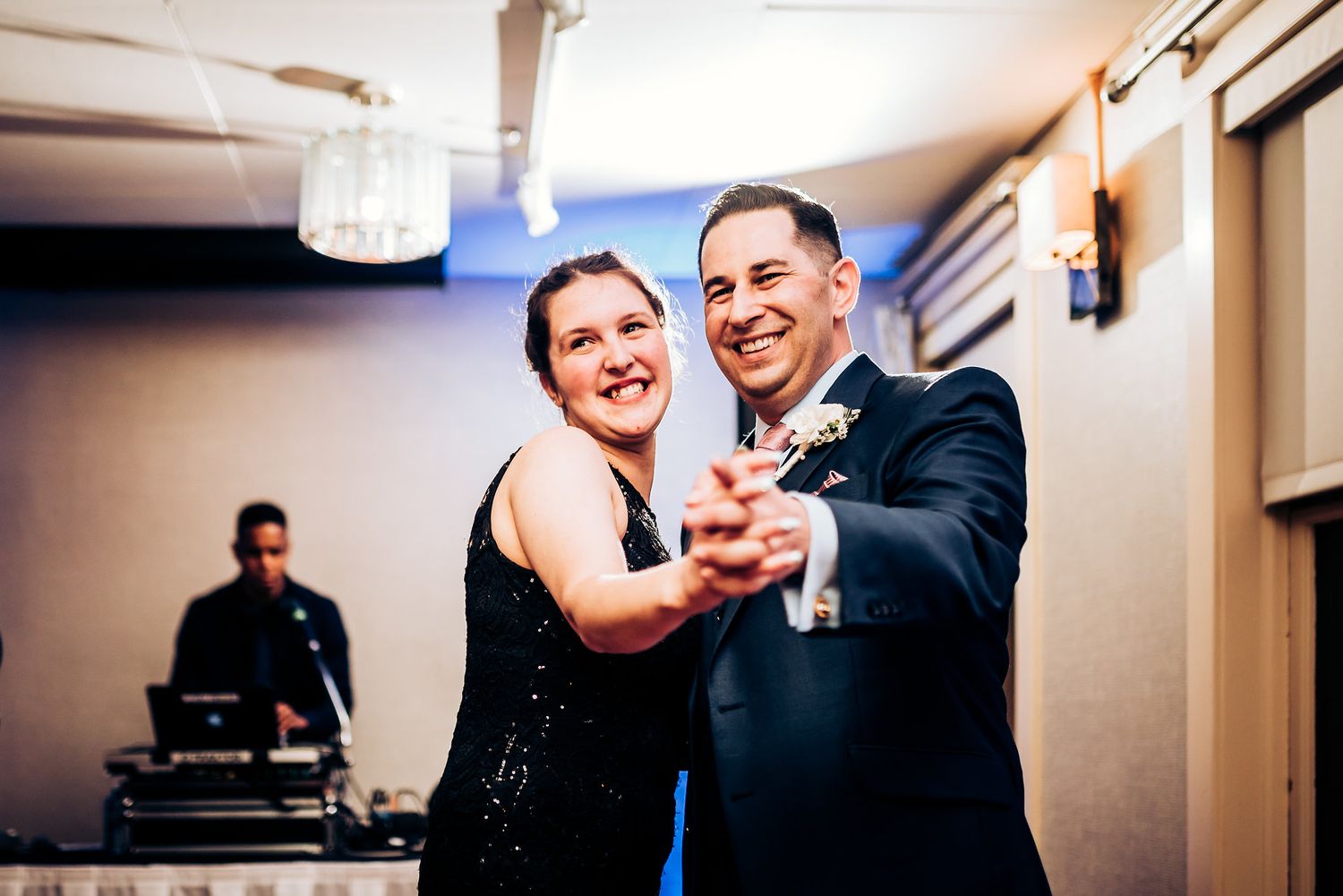 A couple dances together at an indoor reception with a DJ booth visible in the background.