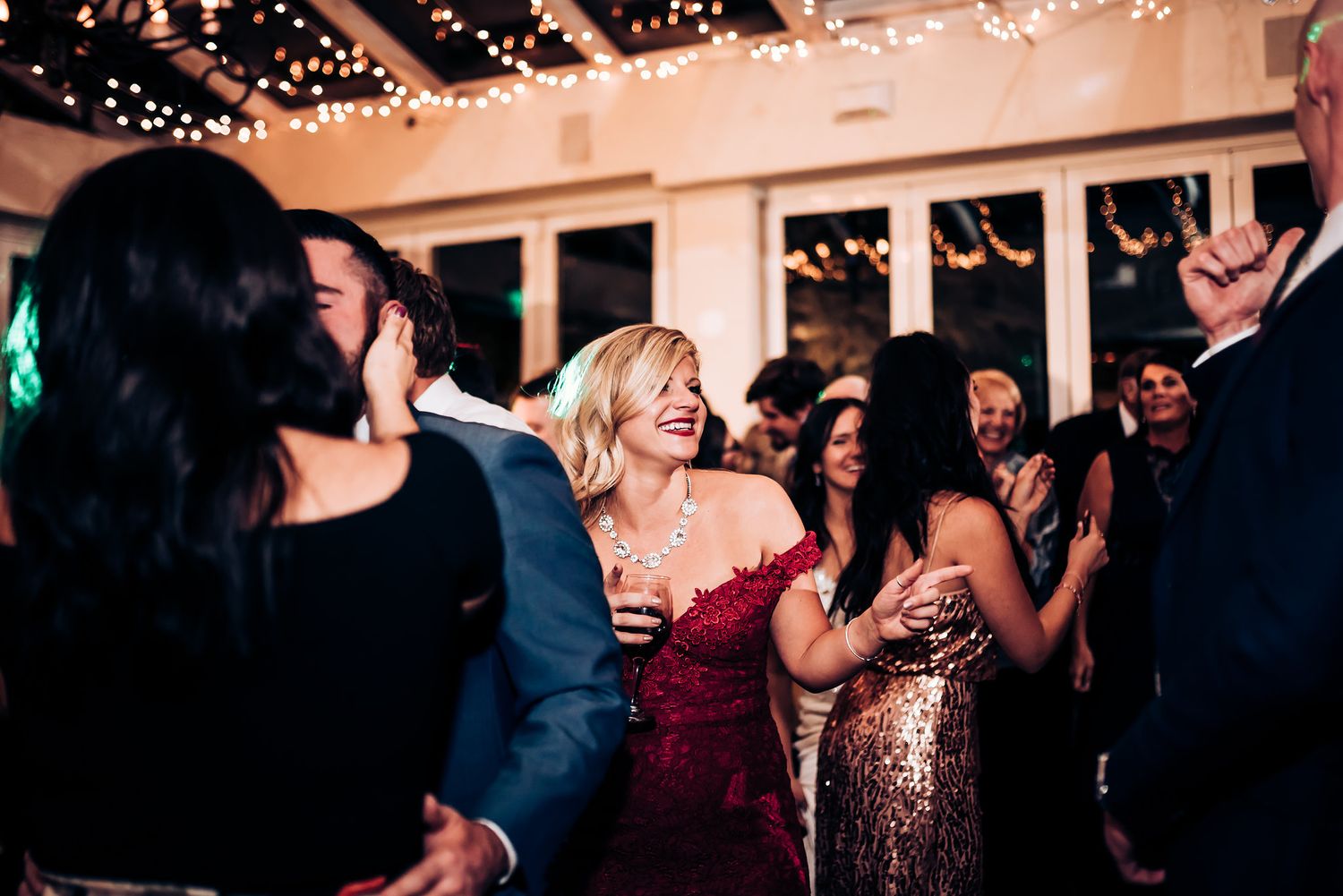 Party guests in formal attire dance and celebrate under twinkling string lights at an indoor reception venue.
