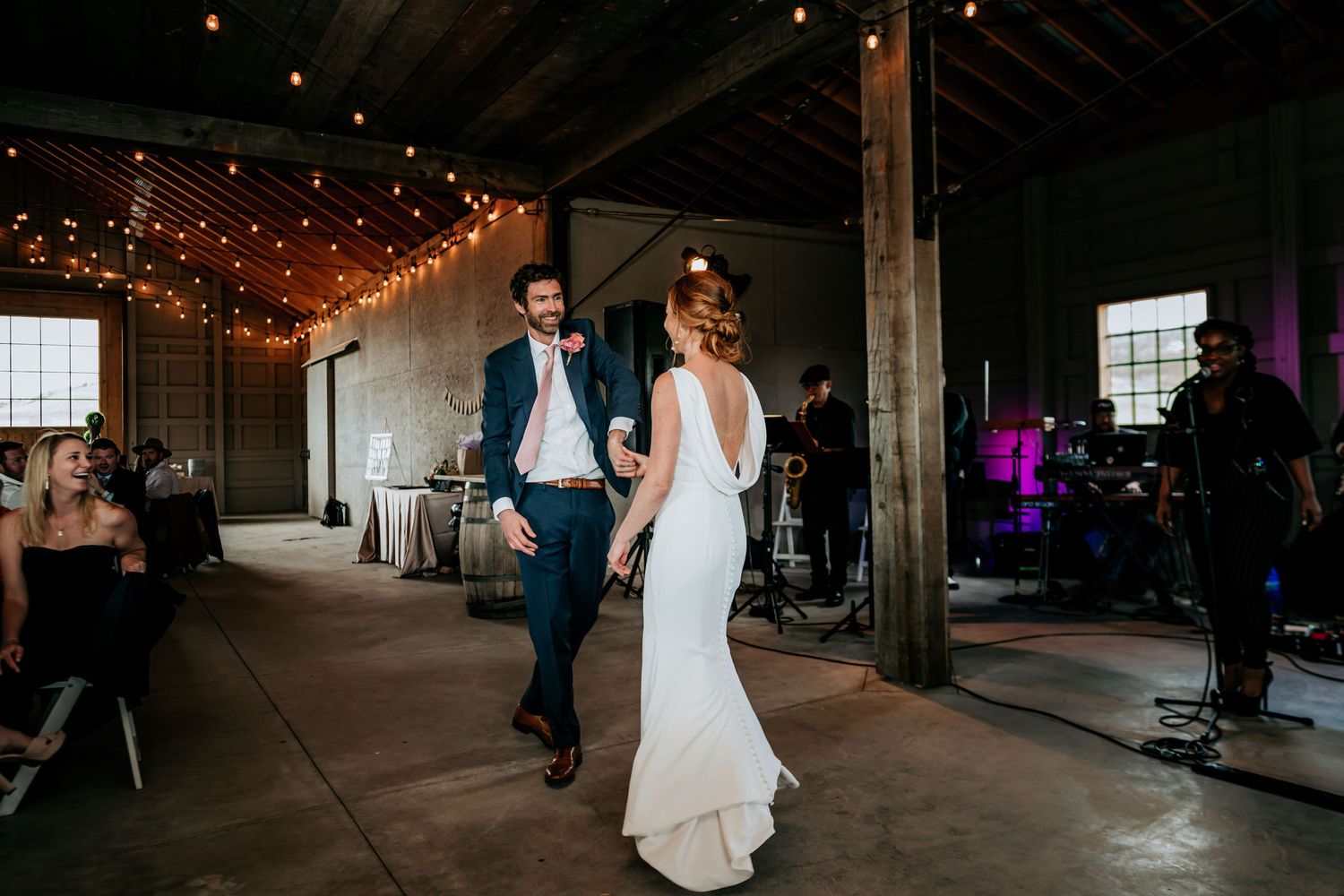 A couple dances in a rustic venue with exposed beams and warm string lights overhead.