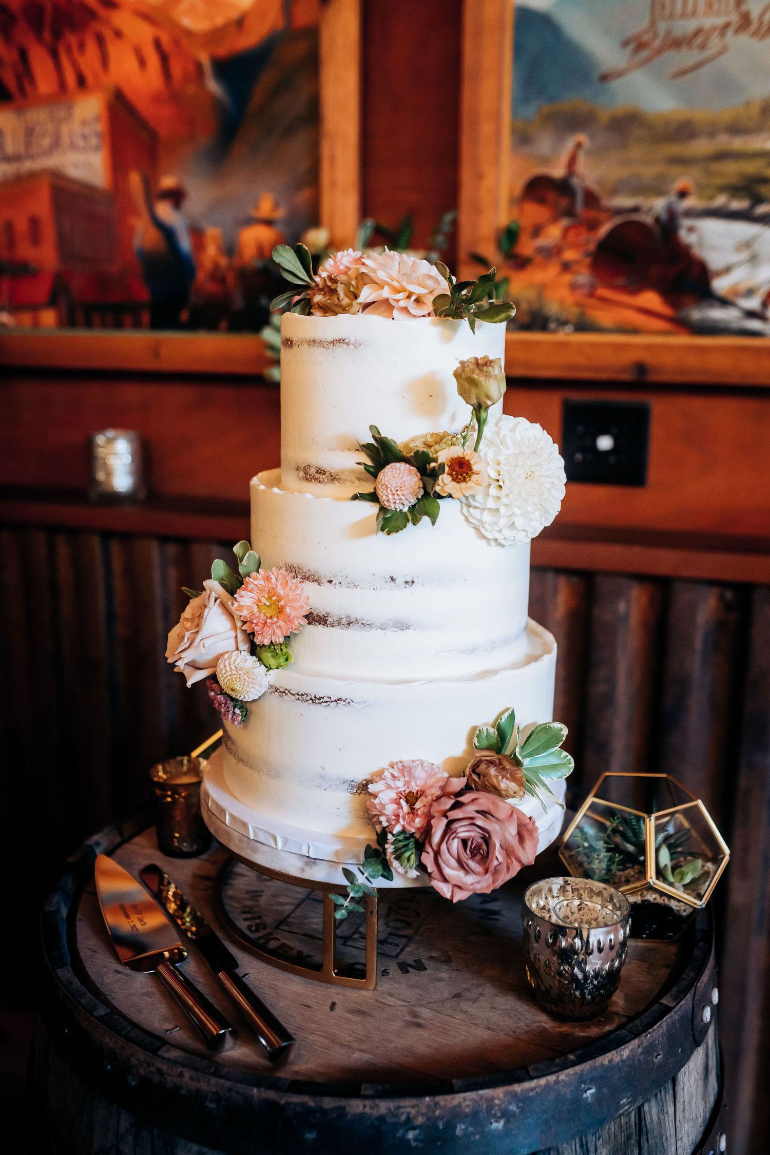 Elegant three-tier white wedding cake decorated with blush roses and greenery on rustic wooden stand.