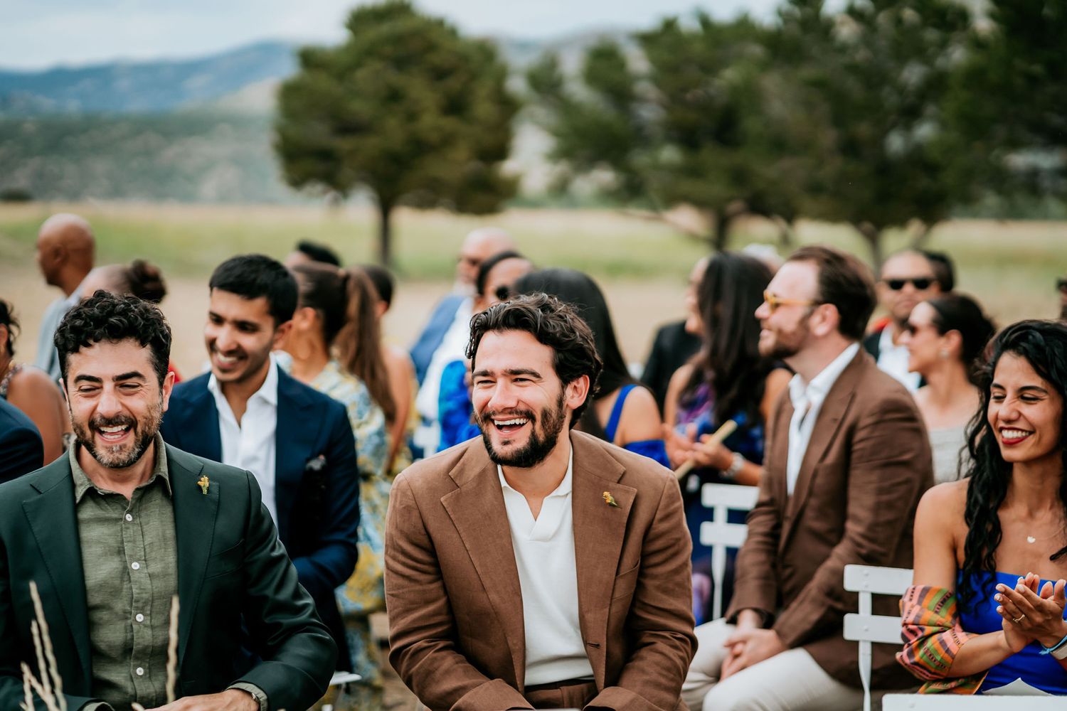 Wedding guests laugh and enjoy themselves during outdoor ceremony in natural setting.