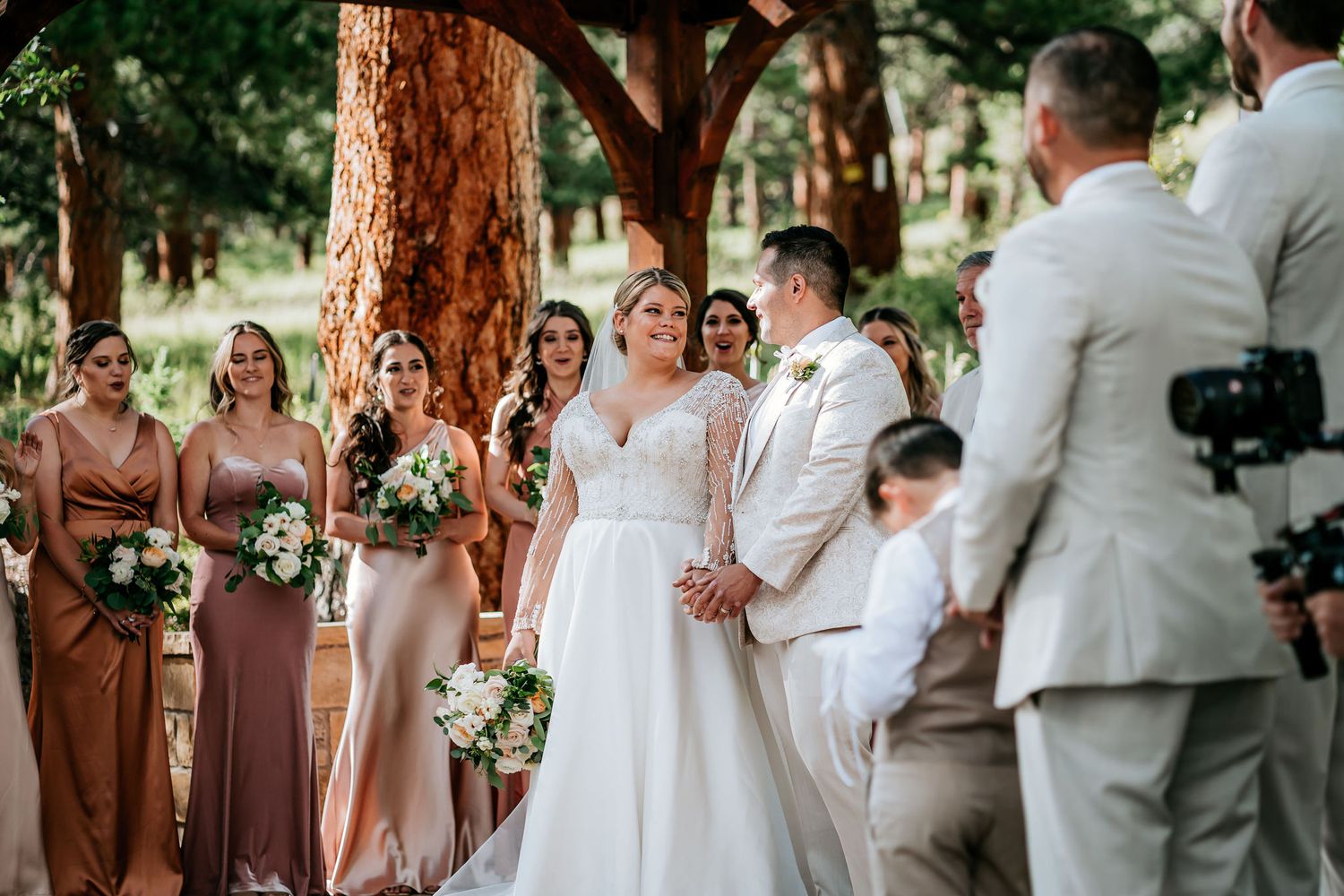 Wedding party in light-colored attire gathered under trees during an outdoor ceremony.