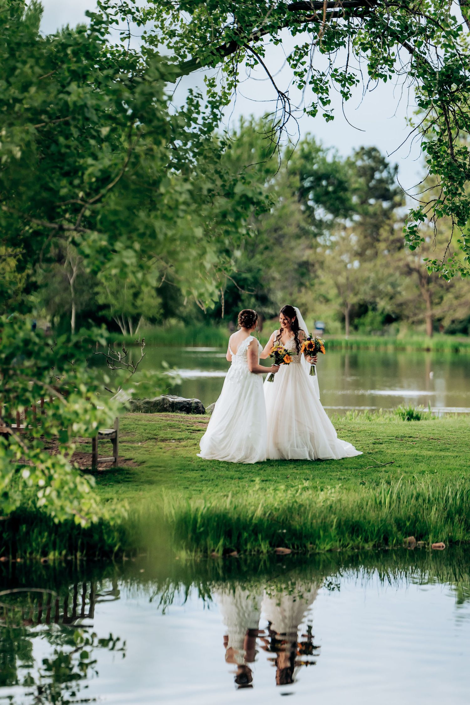 Two brides in white dresses pose by a reflective pond in a lush garden setting.