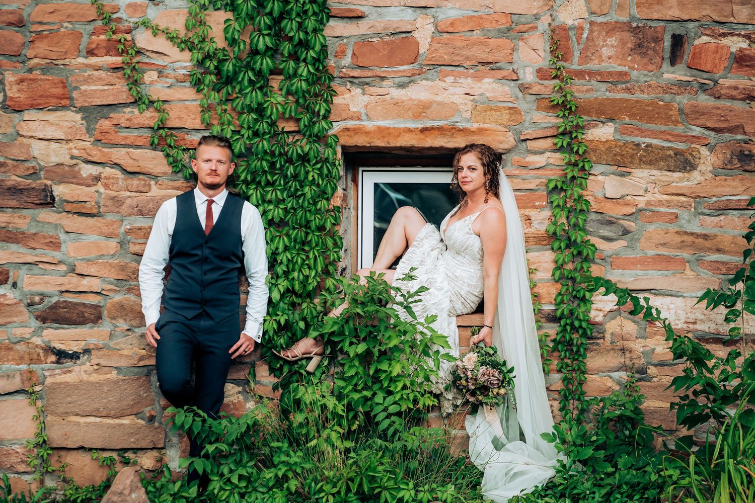A couple poses against an ivy-covered stone wall at a rustic wedding venue.