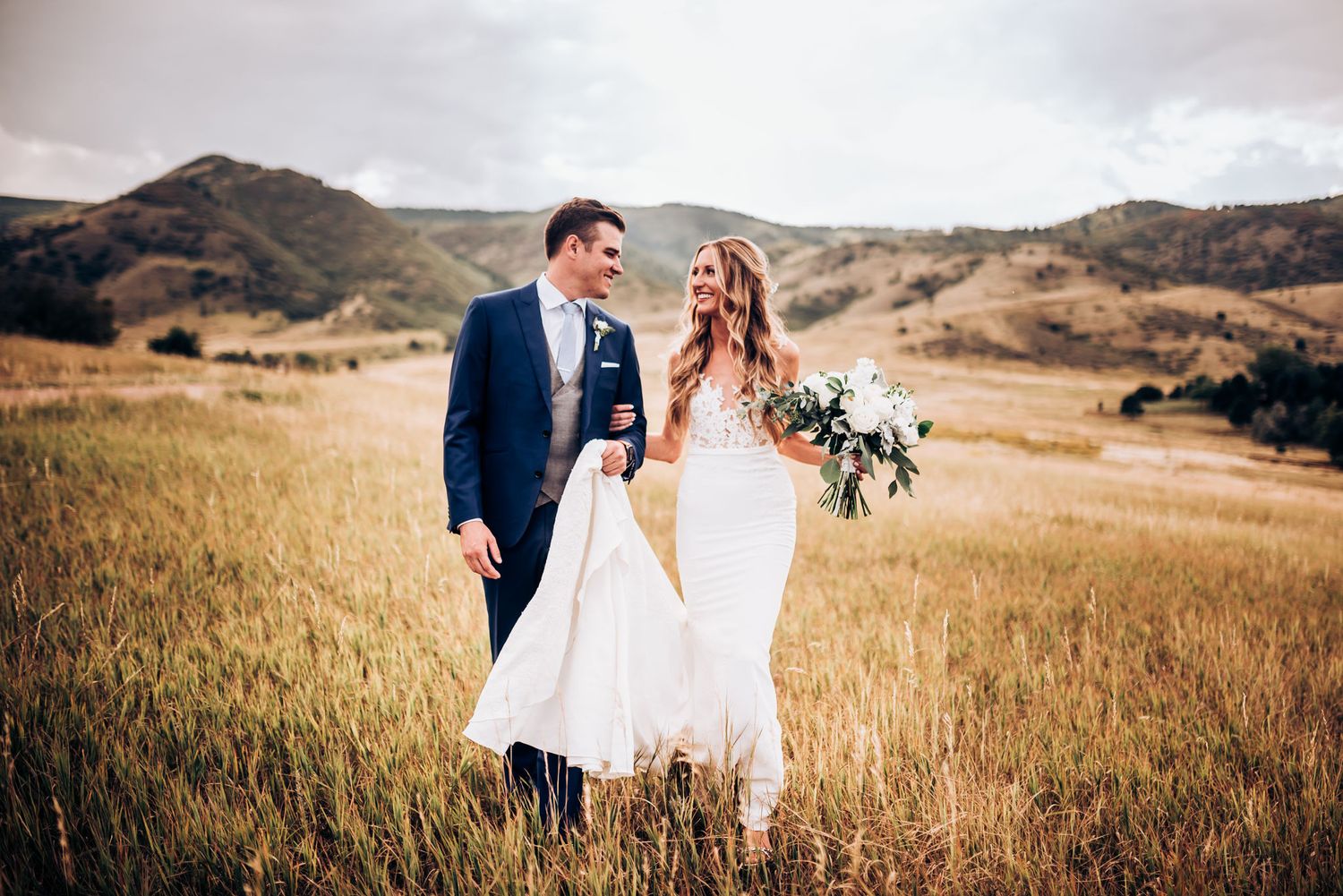 Wedding photo in mountain field with couple holding bouquet against dramatic landscape.
