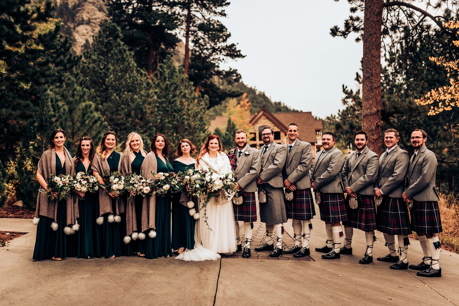 Large wedding party in kilts and formal dresses lined up outdoors at mountain venue.