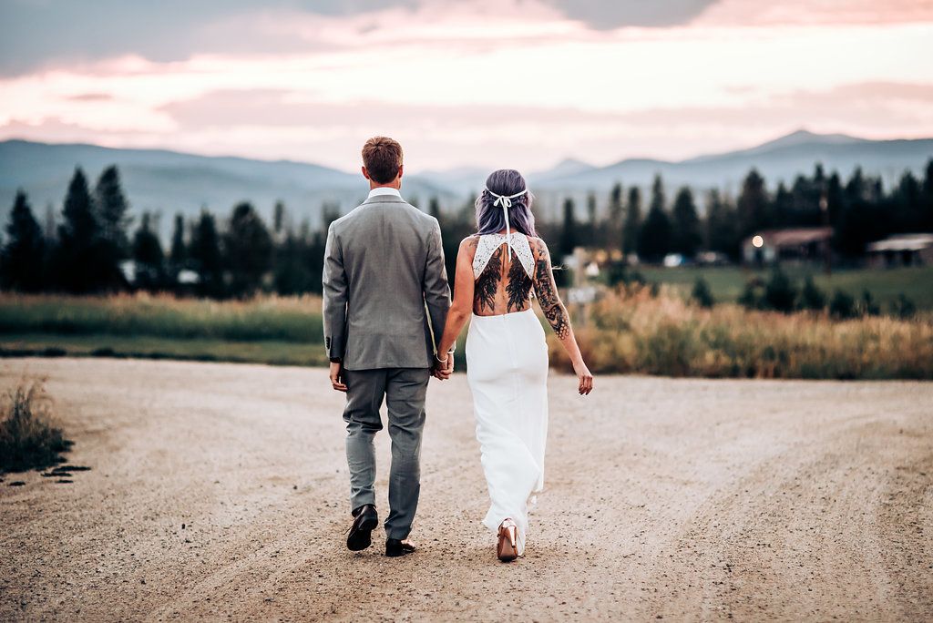 A couple walking down a dirt road with mountain views and pine trees in the background at sunset.