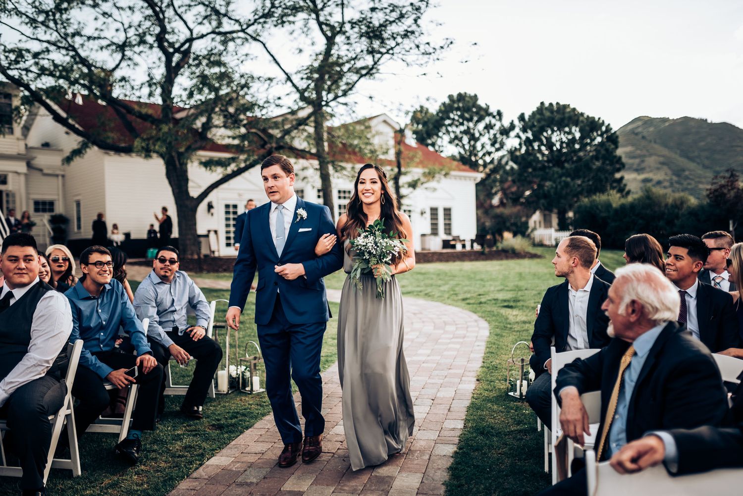 Wedding processional on brick path with guests seated on white chairs near colonial building.