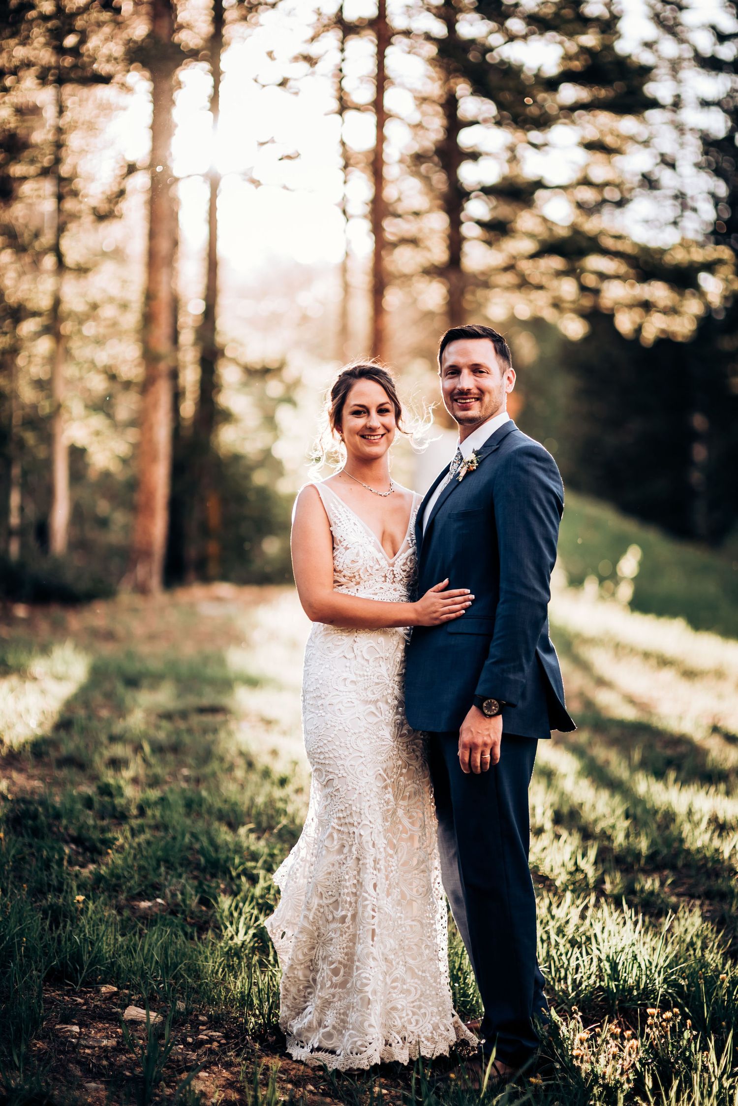 A romantic wedding photo taken in a sunlit forest with a lace dress and navy suit.