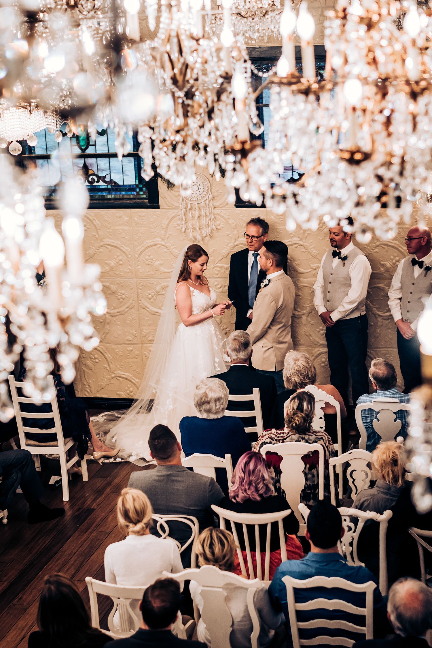 Intimate wedding ceremony with crystal chandeliers and guests seated in antique white chairs.