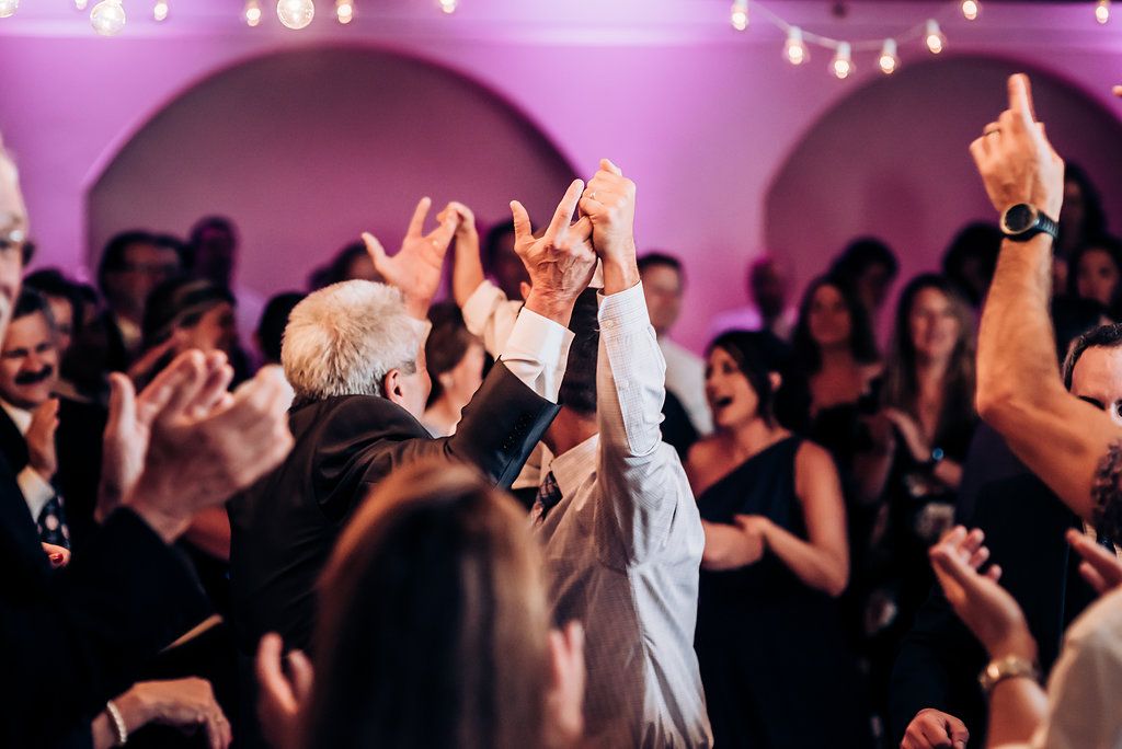 Silhouettes of party guests with raised arms against purple lighting at a celebration.