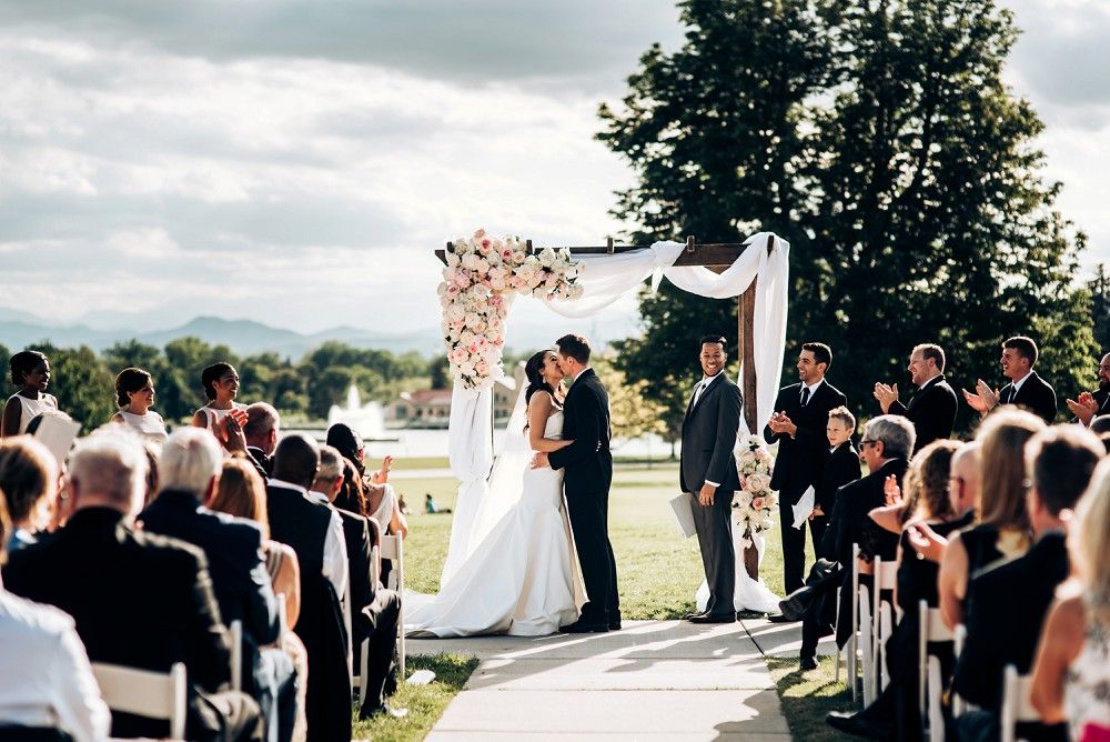 Wedding ceremony under a draped arch with guests seated on white chairs against a cloudy sky.